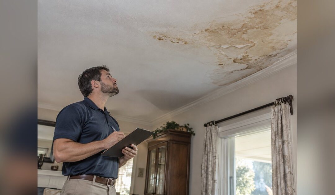 Insurance adjuster in a polo shirt writing on a clipboard while looking up at a water stained ceiling in a Florida living room