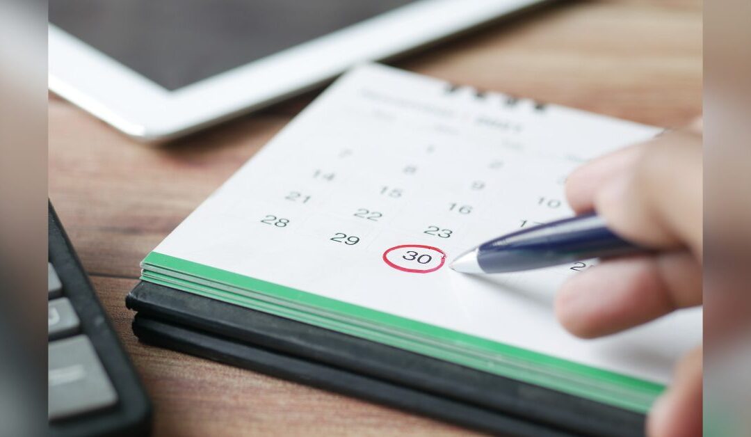 Hand pointing a pen at a date circled in red on a desk calendar